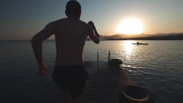 Joven hombre hipster auténtico milenario corre rápido en el paseo marítimo de madera en increíble c natural, lago alpino de montaña con palabra limpia y fresca. salta en forma de bomba divertida. Obtiene placer del viaje de verano . — Vídeo de stock