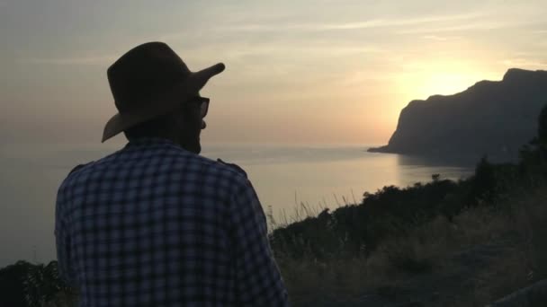 Mladík s kloboukem pozoruje západ slunce na moři slunce zapadá do moře za kapkou. Obdivuje letní cestování. Šťastné chvíle na cestách — Stock video