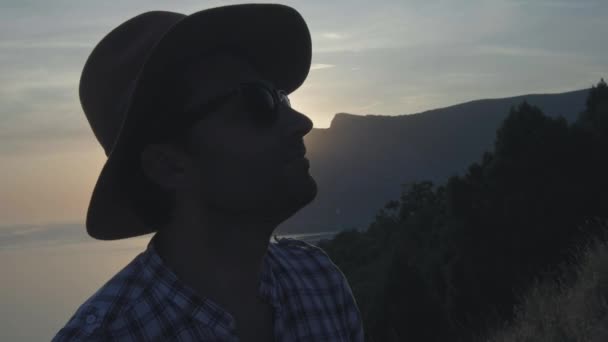 帽子をかぶった若い男が海の夕日を見ている 岬の後ろの海に太陽が沈む。夏の旅行を賞賛します。旅行の幸せな瞬間 — ストック動画