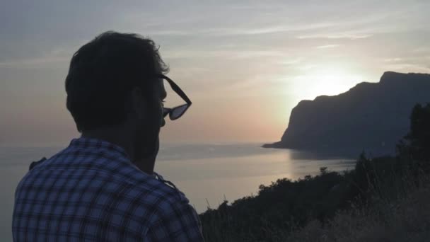 Um jovem com chapéu está a ver o pôr-do-sol no mar. O sol põe-se no mar atrás da capa. Admira viagens de verão. Momentos felizes na viagem — Vídeo de Stock