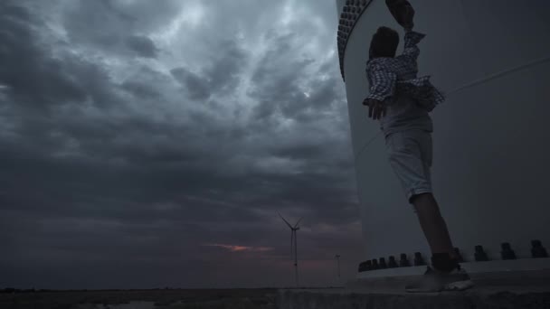 Een jonge kerel naast de wind turbines, produceren ecologische energie. Het concept van milieuvervuiling, nieuwe technologieën van alternatieve energie. Algemeen plan, avond, bewolkt. — Stockvideo