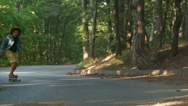 Ein junger Mann lernt, ein Longboard zu fahren. Es liegt im Wald. bekleidet mit kariertem Hemd und Filzhut. Aktiver Lebensstil. — Stockvideo
