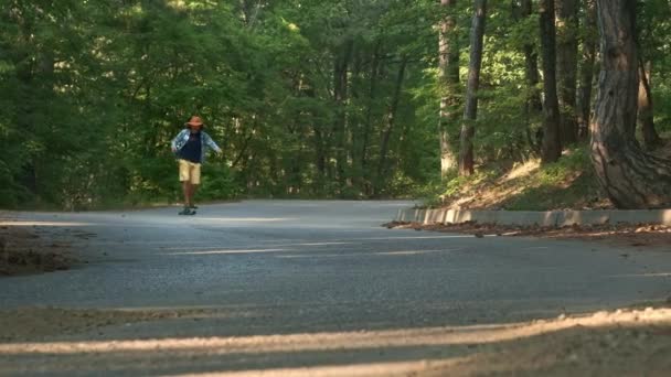 Un joven aprende a montar un longboard. Se encuentra en el bosque. Vestido con una camisa a cuadros y un sombrero de fieltro. Estilo de vida activo . — Vídeo de stock