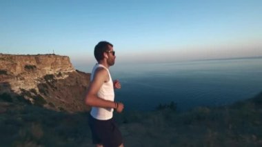 Athletic Handsome Man Telefon ve Kulaklık ile Spor yapıyor, Yaz plajda Jogging sırasında Günbatımında Sea Coast boyunca Çalışan. Kablosuz kulaklık kullanır.