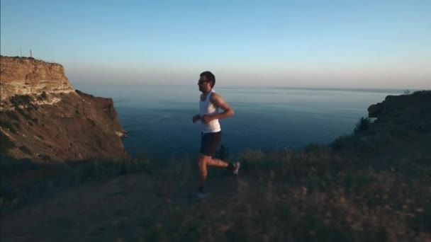 Der athletische, gutaussehende Mann treibt Sport mit Telefon und Kopfhörern und läuft bei Sonnenuntergang an der Küste entlang, während er im Sommer am Strand joggt. verwendet drahtlose Kopfhörer. — Stockvideo