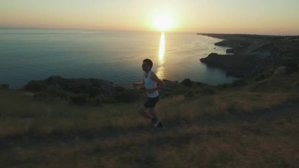 アスレチックハンサムな男は、夏にビーチでジョギングしながら、サンセットで海の海岸に沿って実行し、携帯電話とヘッドフォンでスポーツをしています。ワイヤレスヘッドフォンを使用. — ストック動画