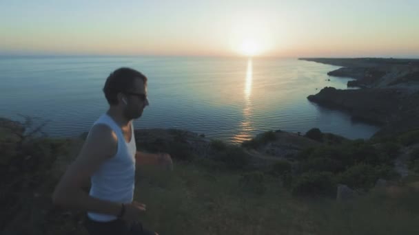 Der athletische, gutaussehende Mann treibt Sport mit Telefon und Kopfhörern und läuft bei Sonnenuntergang an der Küste entlang, während er im Sommer am Strand joggt. verwendet drahtlose Kopfhörer. — Stockvideo