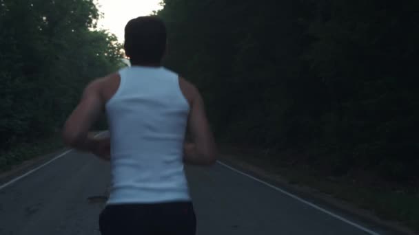 Мужчина выбегает на улицу в дождливый вечер. Мужчина-бегун бегает в сумерках по шоссе среди сосен в лесу. Ведет активный спортивный образ жизни. Укрепляет твое тело. Спорт в природе — стоковое видео