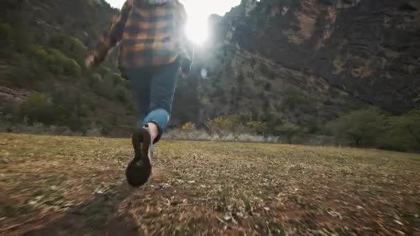 Bella giovane viaggiatrice corre liberamente attraverso il campo nella natura tra le montagne. Lancia un cappello in aria. Gode della vita, della libertà e dei viaggi. Indossare una camicia e un cappello a quadri — Video Stock