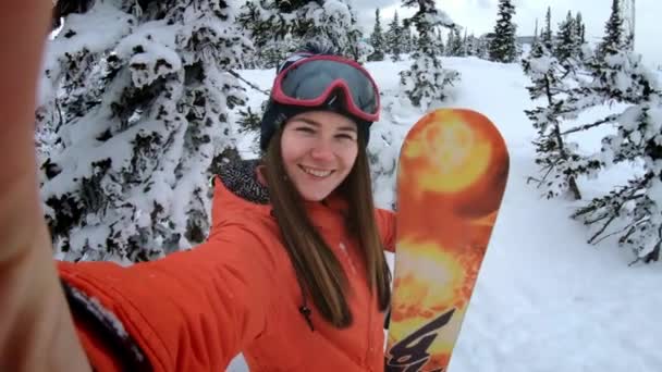 Alegre hermosa niña snowboarder hace selfie en una cámara de acción o teléfono inteligente, sonriendo. Una mujer está de pie entre los pinos nevados de invierno. Diversión invernal en la estación de esquí. Movimiento lento — Vídeo de stock