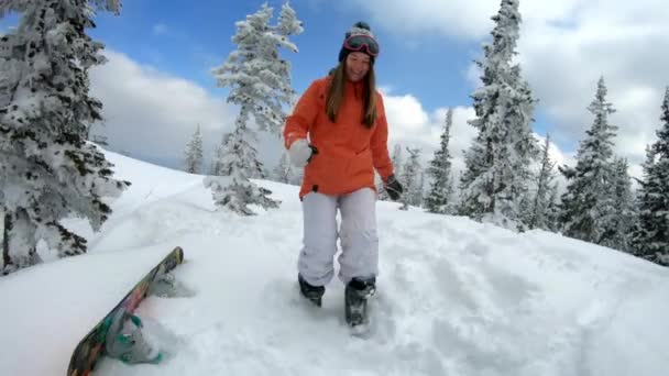Joyeux beau jeune fille snowboarder court dans la neige parmi les pins enneigés. Il tombe à genoux, lance des flocons de neige. Amusement d'hiver à la station de ski. Ralenti, tir de poupée . — Video