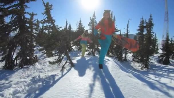 Due belle ragazze allegre snowboarder o sciatori godono seduti in un cumulo di neve e gettando neve, sorridendo. Le donne stanno tra i pini innevati invernali. Divertimento invernale nella stazione sciistica. Rallentatore — Video Stock