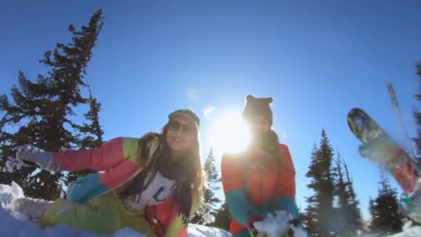 Zwei fröhlich schöne junge Mädchen Snowboarder oder Skifahrer genießen es, lächelnd in einer Schneewehe zu sitzen und Schnee zu werfen. Frauen stehen zwischen winterlich schneebedeckten Kiefern. Winterspaß im Skigebiet. Zeitlupe — Stockvideo