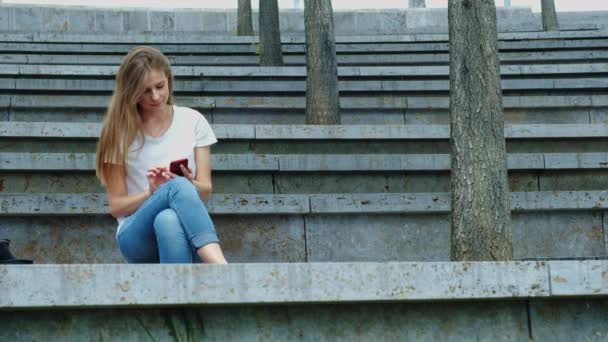 Joven chica hermosa en un parque tiene un teléfono inteligente en sus manos, escribe un mensaje. Hojeando fotos por teléfono, charlando en redes sociales con amigos. Se sienta sobre hormigón — Vídeos de Stock