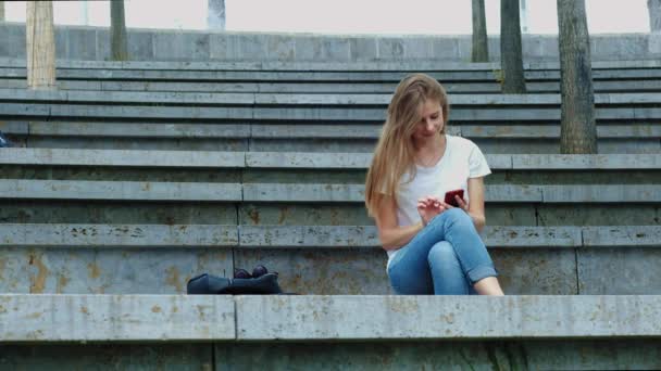 Jong mooi meisje in een park houdt een smartphone in haar handen, schrijft een bericht. Bladeren door Foto's op de telefoon, chatten in sociale netwerken met vrienden. Zit op beton — Stockvideo