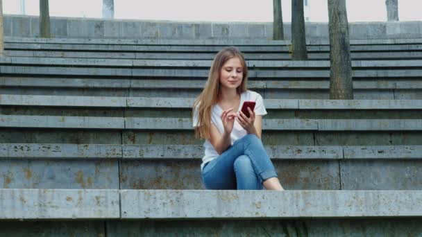 Young beautiful girl in a park holds a smartphone in her hands, writes a message. Leafing through photos on the phone, chatting in social networks with friends. Sits on concrete — Stock Video