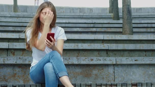Young beautiful girl in a park holds a smartphone in her hands, writes a message. Leafing through photos on the phone, chatting in social networks with friends. Sits on concrete — Stock Video