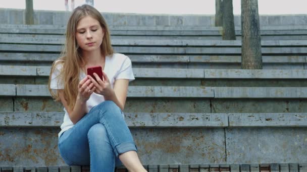 Młoda piękna dziewczyna w parku trzyma smartfon w dłoniach, pisze wiadomość. Leafing przez zdjęcia w telefonie, rozmowy w sieciach społecznościowych z przyjaciółmi. Siedzi na betonie — Wideo stockowe