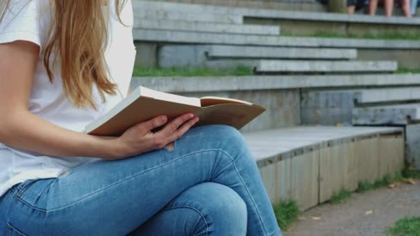 Ung vacker flicka i en Park läser en bok. En elev studerar en bok, svarvning sidor. Sitter på betong — Stockvideo