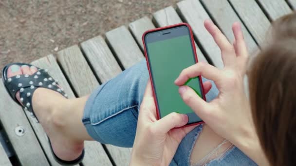 Ένα νεαρό όμορφο κορίτσι στο πάρκο. Κρατά στα χέρια του ένα μαύρο smartphone με πράσινη οθόνη. Το θηλυκό χέρι κρατά ένα τηλέφωνο με χρώμιο Κέι. Κάνει το ζουμ με δύο δάκτυλα, εξετάζει τον χάρτη — Αρχείο Βίντεο