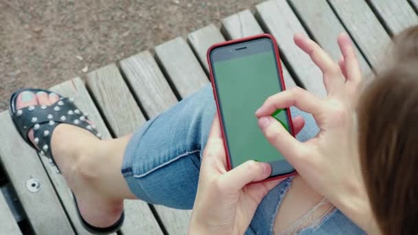 Jeune belle fille dans le parc. Il tient dans ses mains un smartphone noir avec un écran vert. Main féminine tient un téléphone avec chrome kay. Fait le zoom avec deux doigts, examine la carte — Video