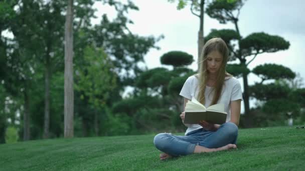 Młoda piękna dziewczyna w parku czyta książkę. Uczeń studiuje książkę, obracając strony. Siedzi na trawie, na tle pięknych drzew. — Wideo stockowe