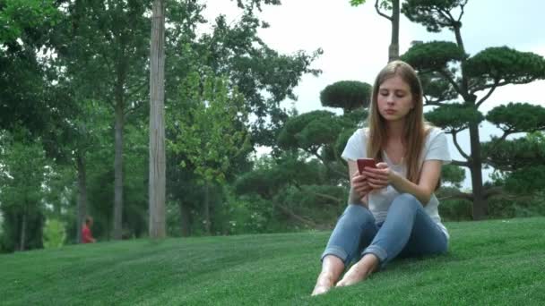 公园里漂亮的年轻女孩在智能手机上自拍，给她的男朋友发一张照片。翻阅手机上的照片，在社交网络上与朋友聊天。坐在草地上. — 图库视频影像