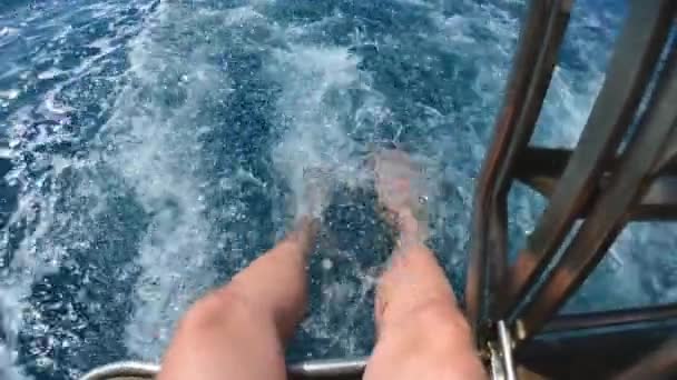 Um jovem bonito senta-se com as pernas para baixo de um barco ou iate no mar ou no oceano. Brinca com água, spray voador. Férias de verão com amigos. Movimento lento . — Vídeo de Stock