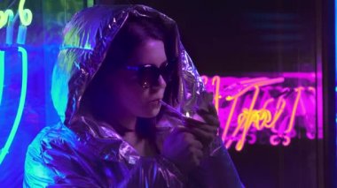 Bin yıllık serin modern güzel kız geceleri parlayan neon duvar yakınında sigara içiyor. Gizemli hipster. Parlak lazer ceket ve güneş gözlüğü giyen güzel şık genç. kötü yaşam tarzı.