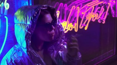 Bin yıllık serin modern güzel kız geceleri parlayan neon duvar yakınında sigara içiyor. Gizemli hipster. Parlak lazer ceket ve güneş gözlüğü giyen güzel şık genç. kötü yaşam tarzı.