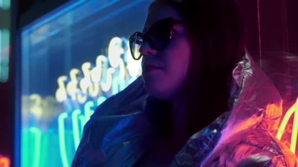 年轻时尚少女在毛皮眼镜吹泡泡糖照明与街头霓虹灯蓝色粉红色标志，美丽的千禧年妇女在时髦的夜光辉回80年代的概念 — 图库视频影像