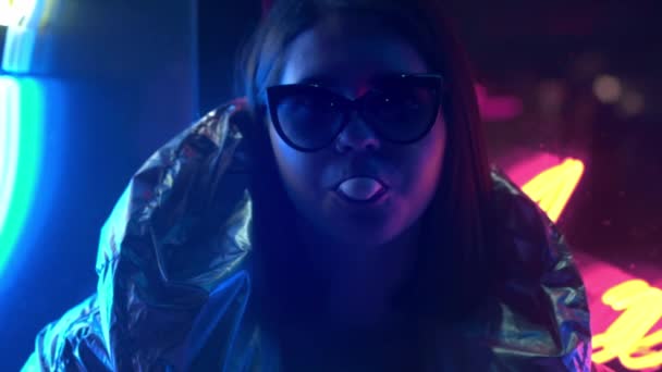 Young fashion teen girl in fur glasses blowing bubble gum illuminated with street neon blue pink sign, beautiful millennial woman in trendy night light glow back to 80s concept — Stock Video