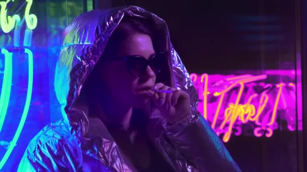 Millennial cool modern pretty girl smoking cigarette near glowing neon wall at night. Mysterious hipster. Beautiful stylish teen wearing shiny laser jacket and sunglasses. bad lifestyle. — Stock Video
