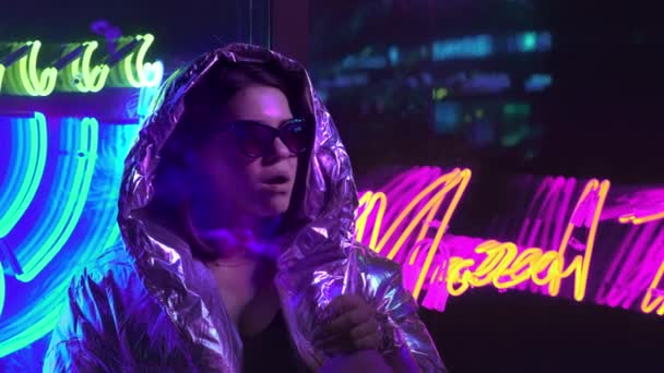 Millennial cool modern Pretty Girl röka cigarett nära glödande Neon Wall på natten. Mystiska hipster. Vacker snygg tonåring klädd i glänsande laser jacka och solglasögon. dålig livsstil. — Stockvideo