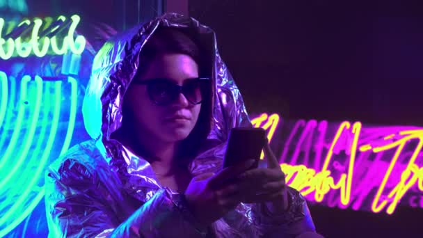 Ung flicka Messaging i mobiltelefon Chat App. mörk plats med blandat färgat ljus. Kommunikation sociala medier koncept. Vacker snygg tonåring klädd i glänsande laser jacka och solglasögon. — Stockvideo