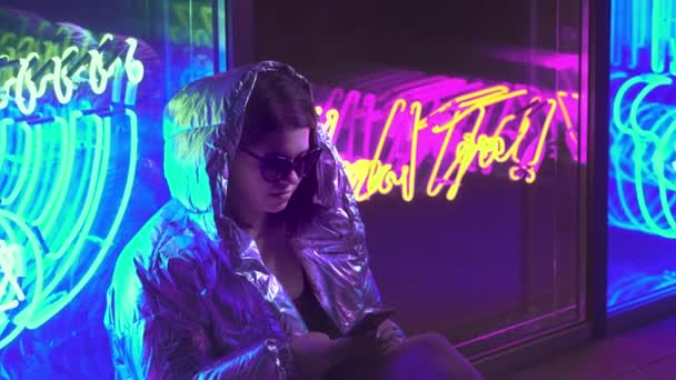Young girl messaging in cellphone chat app. Dark place with mixed coloured light. Communication social media concept. Beautiful stylish teen wearing shiny laser jacket and sunglasses. — Stock Footage Young girl messaging in cellphone chat app. Dark place with mixed coloured light. Communication social media concept. Beautiful stylish teen wearing shiny laser jacket and sunglasses. — Stock Video