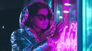 Geceleri parlayan neon duvarın yanında bin yıllık esrarengiz güzel kız. Gözlüklü gizemli hipster genç. Neon Lights'a dokunmak. Parlak lazer ceket ve güneş gözlüğü giyen güzel şık genç