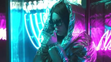 Bin yıllık serin modern güzel kız geceleri parlayan neon duvar yakınında sigara içiyor. Gizemli hipster. Parlak lazer ceket ve güneş gözlüğü giyen güzel şık genç. kötü yaşam tarzı.