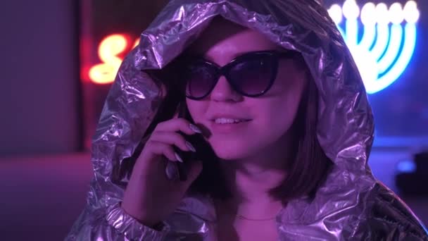 Chica bonita atractiva cerca de luces de neón brillantes de la ciudad por la noche hablando con el teléfono inteligente. Feliz adolescente hipster usando el móvil. Hermosa adolescente elegante con brillante chaqueta láser y gafas de sol — Vídeos de Stock