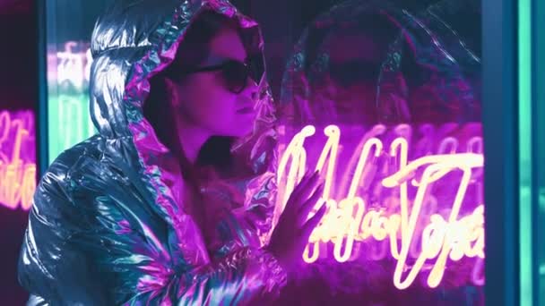Millennial enigmatic pretty girl near glowing neon wall at night. Mysterious hipster teenager in glasses. Touching Neon Lights. Beautiful stylish teen wearing shiny laser jacket and sunglasses — Stock Video
