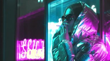 Bin yıllık serin modern güzel kız geceleri parlayan neon duvar yakınında sigara içiyor. Gizemli hipster. Parlak lazer ceket ve güneş gözlüğü giyen güzel şık genç. kötü yaşam tarzı.