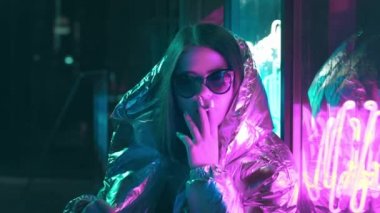 Bin yıllık serin modern güzel kız geceleri parlayan neon duvar yakınında sigara içiyor. Gizemli hipster. Parlak lazer ceket ve güneş gözlüğü giyen güzel şık genç. kötü yaşam tarzı.