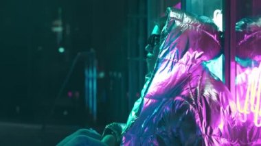 Geceleri parlayan neon duvarın yanında bin yıllık esrarengiz güzel kız. Gözlüklü gizemli hipster genç. Parlak lazer ceket ve güneş gözlüğü giyen güzel şık genç. kötü yaşam tarzı.