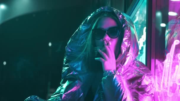 Millennial cool modern Pretty Girl röka cigarett nära glödande Neon Wall på natten. Mystiska hipster. Vacker snygg tonåring klädd i glänsande laser jacka och solglasögon. dålig livsstil. — Stockvideo