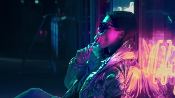 Young fashion teen girl in fur glasses licks a candy illuminated with street neon blue pink sign, beautiful millennial woman in trendy night light glow back to 80s concept. Big lollipop in the mouth. — Stock Video
