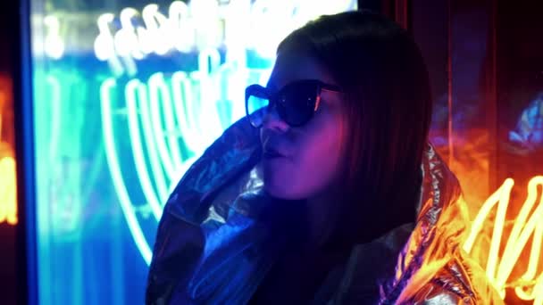 Девушка в меховых очках, надувающая жвачку, освещенную уличным неоновым синим розовым знаком, красивая женщина тысячелетия в модном ночном свете, светящемся назад к концепции 80-х годов — стоковое видео