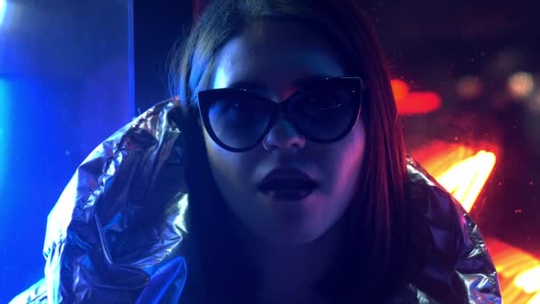 Девушка в меховых очках, надувающая жвачку, освещенную уличным неоновым синим розовым знаком, красивая женщина тысячелетия в модном ночном свете, светящемся назад к концепции 80-х годов — стоковое видео
