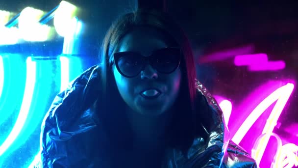 Young fashion teen girl in fur glasses blowing bubble gum illuminated with street neon blue pink sign, beautiful millennial woman in trendy night light glow back to 80s concept — Stock Video