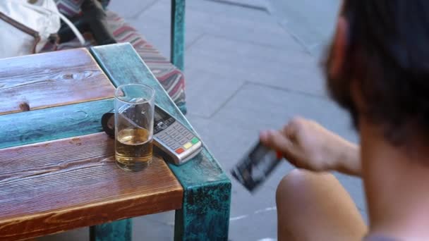 Un joven barbudo viajero o turista en un café o restaurante aplica una tarjeta bancaria con el módulo NFC. Paga con dinero electrónico, tarjeta sin contacto. Utiliza un terminal para el pago sin efectivo — Vídeos de Stock