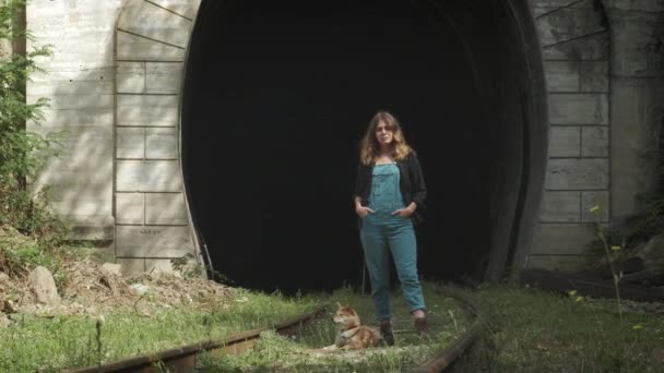 Дівчина стоїть на тлі старого тунелю. Дівчина з собакою стоїть на залізниці — стокове відео